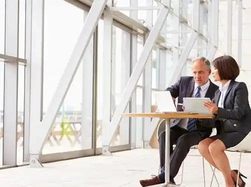 work-smarter_58a5ceebc2f23f7333c95630541e1523 Businessman and businesswoman looking over laptops near window wall.